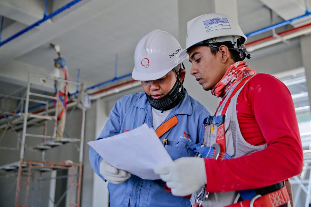 Two electricians wearing safety gear and reviewing construction blueprints on a job site before starting an electrical contracting business.
