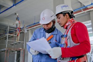 Two electricians wearing safety gear and reviewing construction blueprints on a job site before starting an electrical contracting business.